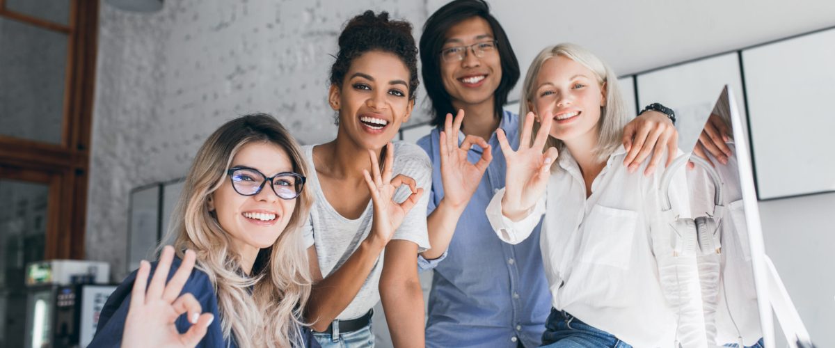 Team of talented young web-developers done with hard project and posing with smile. Asian student in glasses standing beside african cute girl and showing okay sign in office.